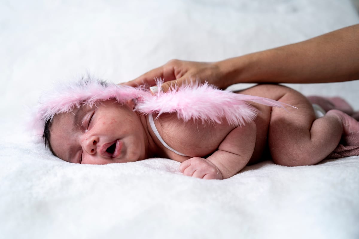 A baby wearing a pink feathered headband, lying on a white blanket.