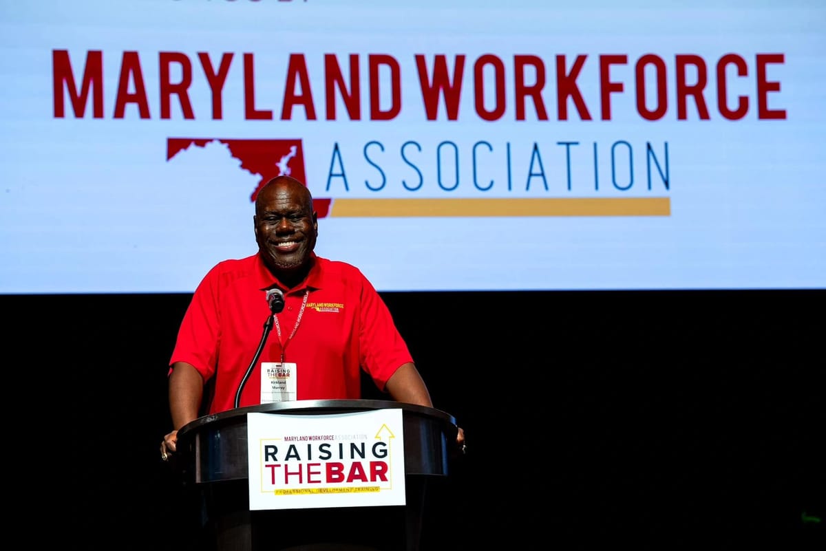 A man in a red polo shirt stands at a podium with a microphone, facing the camera.