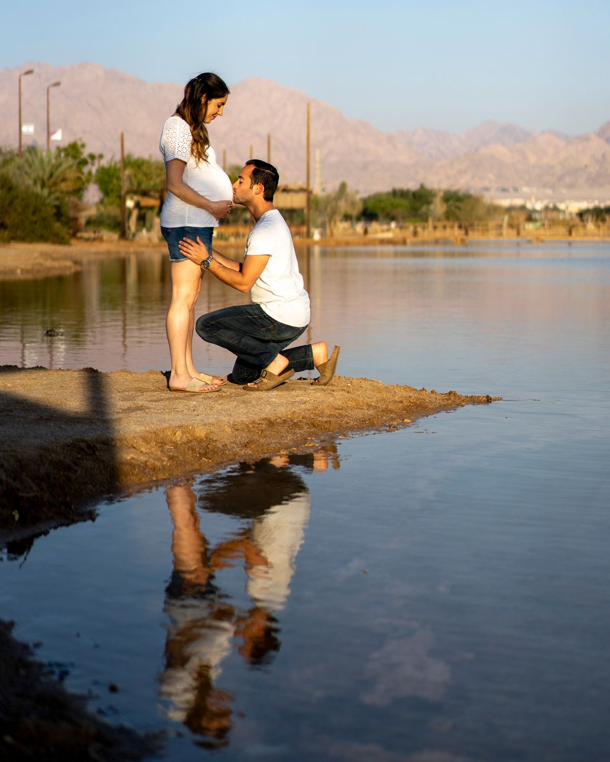 A man and woman, both wearing white t-shirts, stand on a rocky outcropping in a body of water, with a mountain range in the background.