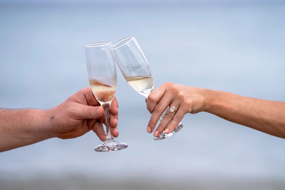 A man and woman's hands clinking champagne flutes together, with the woman's left hand showing an engagement ring.