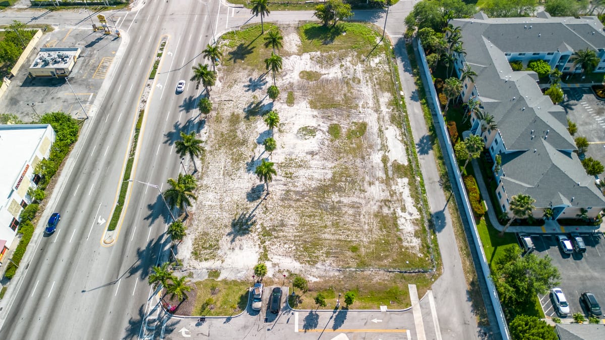 Aerial view of a vacant lot in a suburban area, surrounded by palm trees and a road.