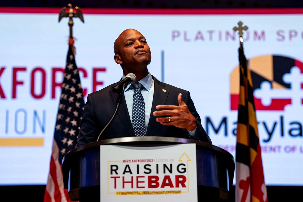A man in a dark suit and blue tie stands at a podium, addressing an audience.