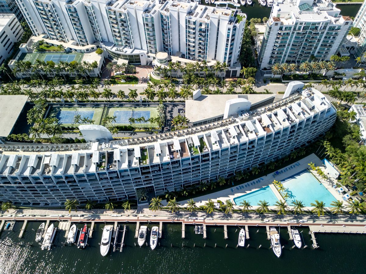 Aerial view of a large white building with a pool and marina, surrounded by palm trees and a docked boat.