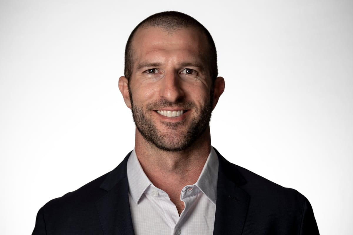 Man in a dark suit jacket and white collared shirt, smiling against a neutral grey backdrop.