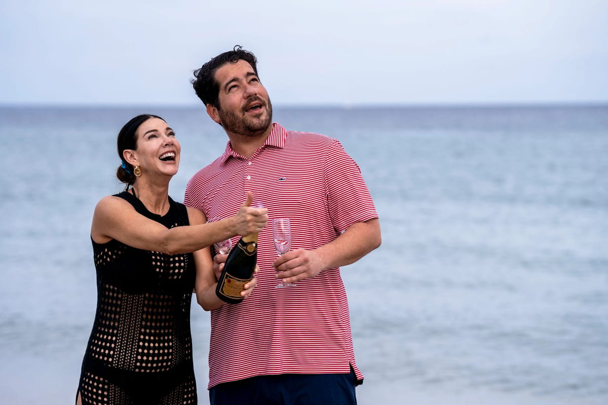 A man and woman standing on a beach, holding champagne flutes and a bottle of champagne, smiling at each other.