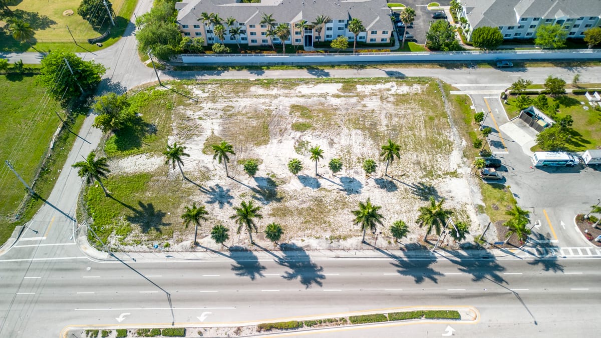 Aerial view of a vacant lot with palm trees and a road in front of it, with buildings in the background.