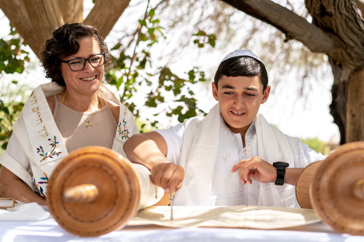 A boy in a white shirt and kippah, smiling at the camera, with a woman in a white dress and shawl standing next to him.