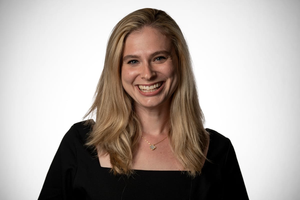 A woman with long blonde hair and a black top smiles at the camera against a white backdrop.