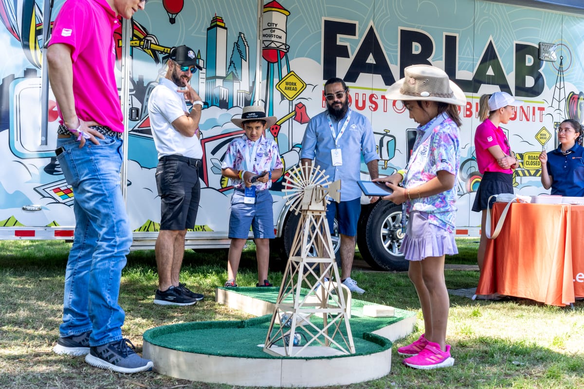 A young girl in a floral shirt and purple skirt stands in front of a miniature golf course, holding a tablet and looking at a model windmill.