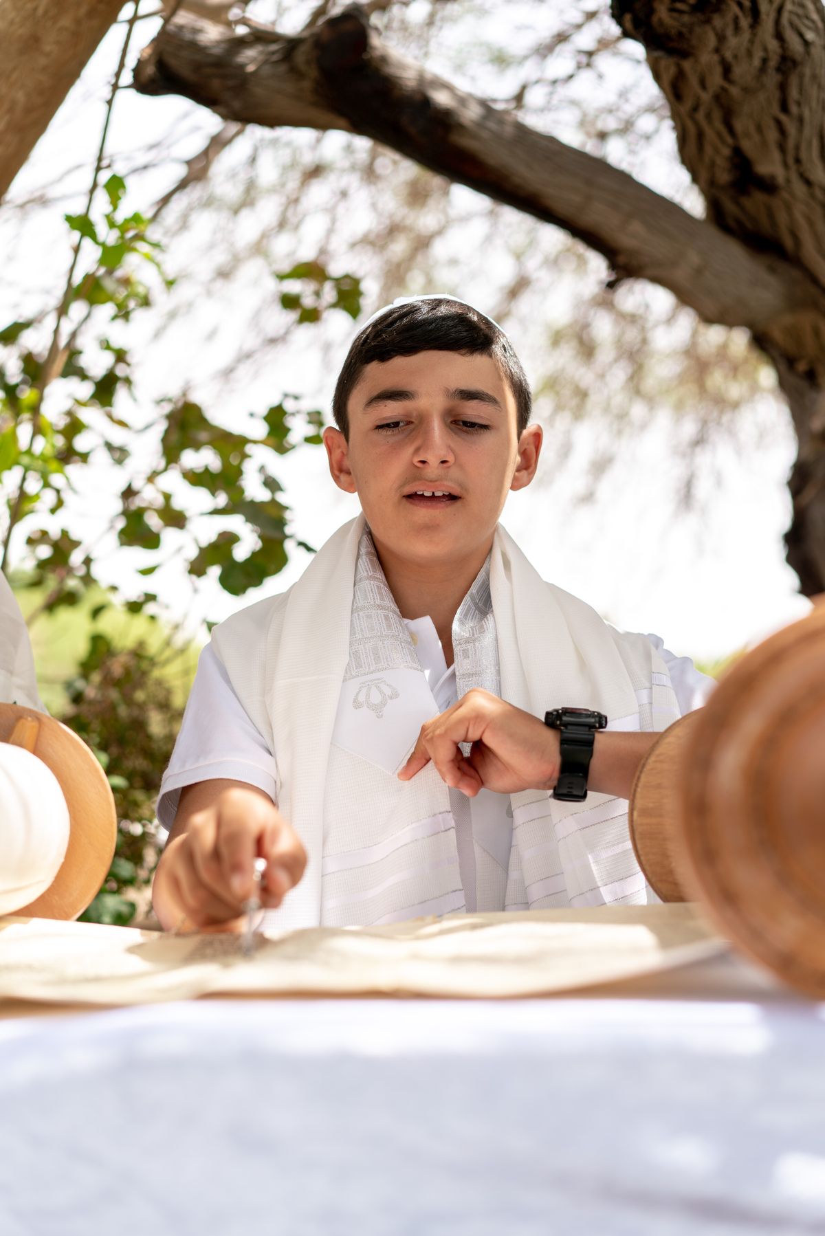 A young boy in a white shirt and tallit, sitting at a table with a white tablecloth, holding a spoon in his right hand.