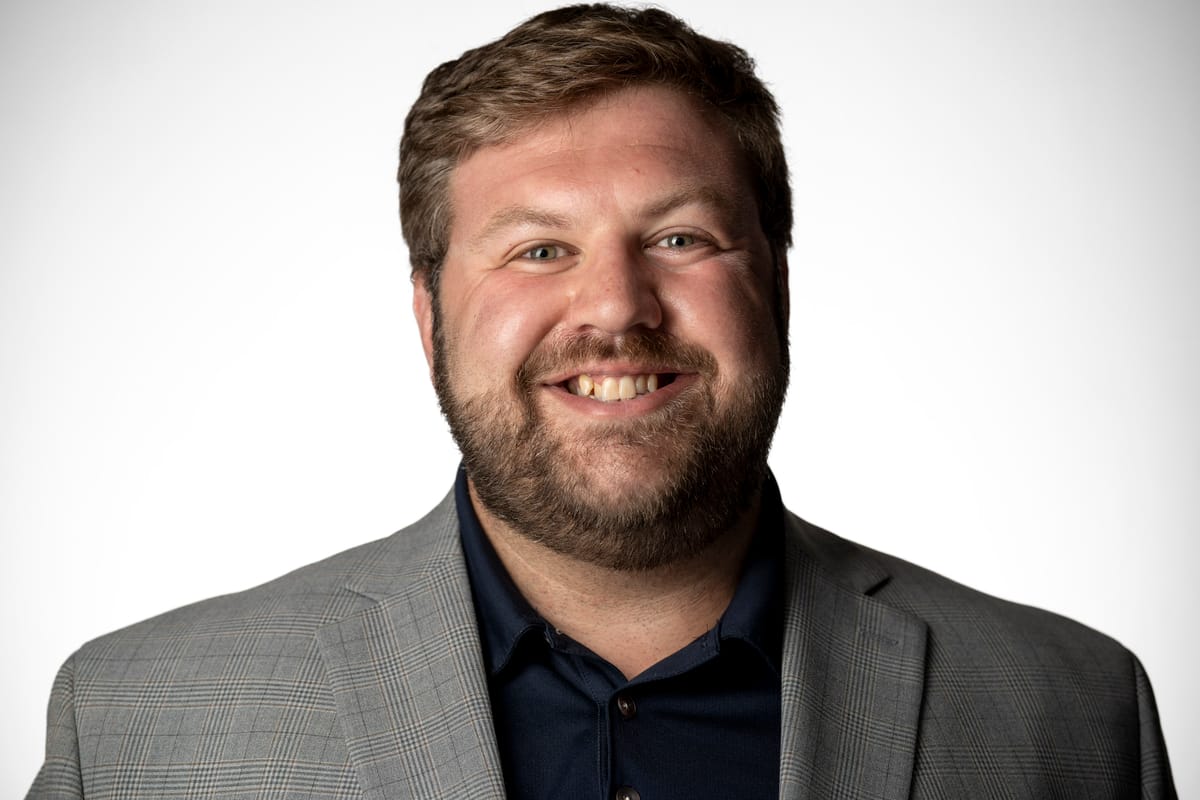 Man in a gray plaid suit jacket and dark blue collared shirt, visible from the chest up, against a plain white backdrop.