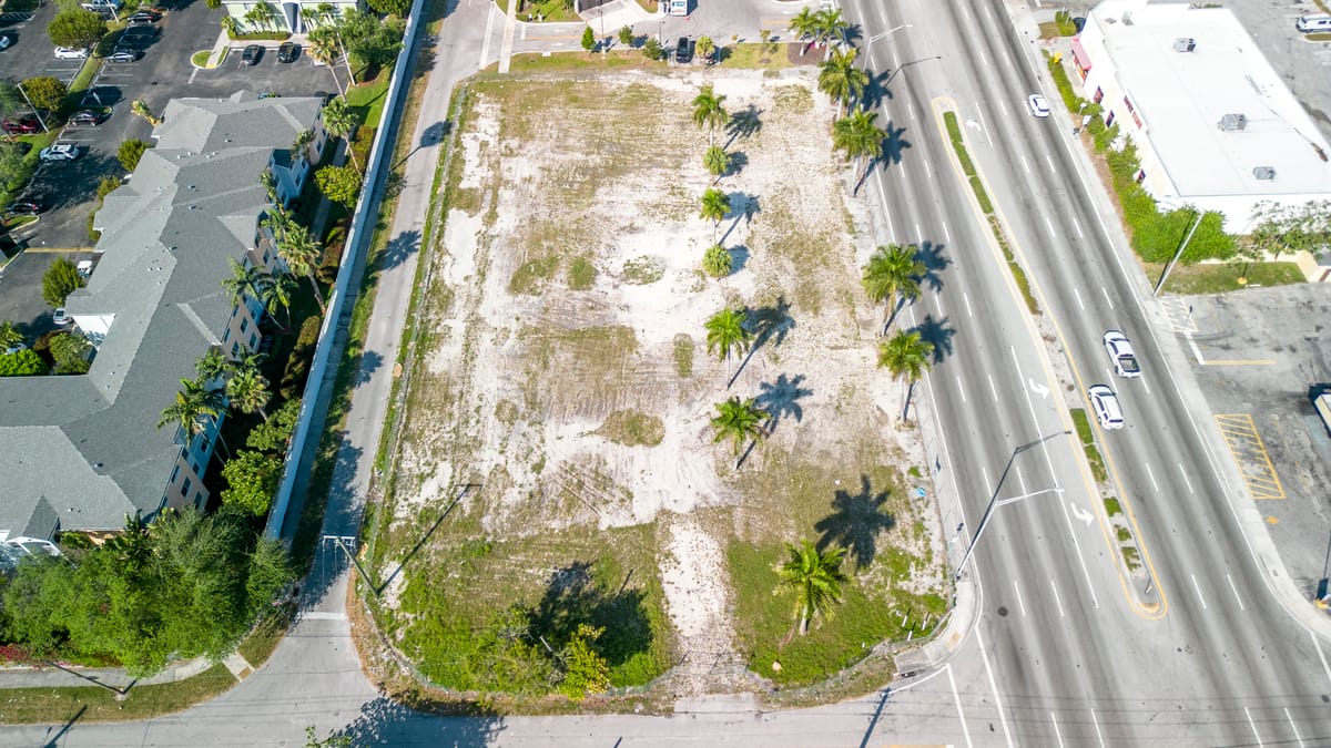Aerial view of a vacant lot in a residential area, surrounded by palm trees and a road.