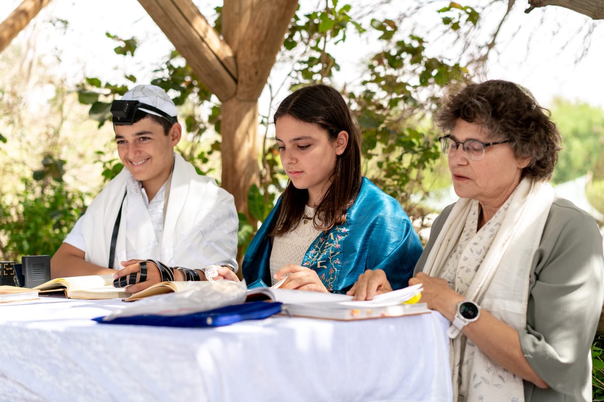 A young girl and boy sitting at a table, surrounded by books and papers, with a woman standing beside them.