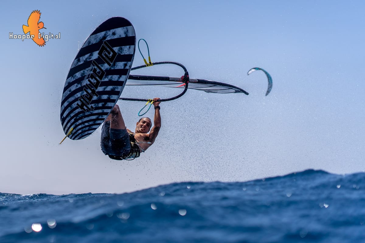 A man in a black and white striped shirt, holding a windsurfing board, in mid-air above the water.