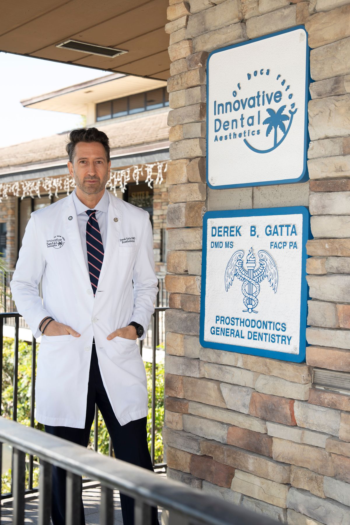 A dentist in a white lab coat stands outside a building with a sign that reads 'Innovative Dental Aesthetics' and 'Derek B. Gatta, DMD MS FACP PA Prosthodontics General Dentistry'.